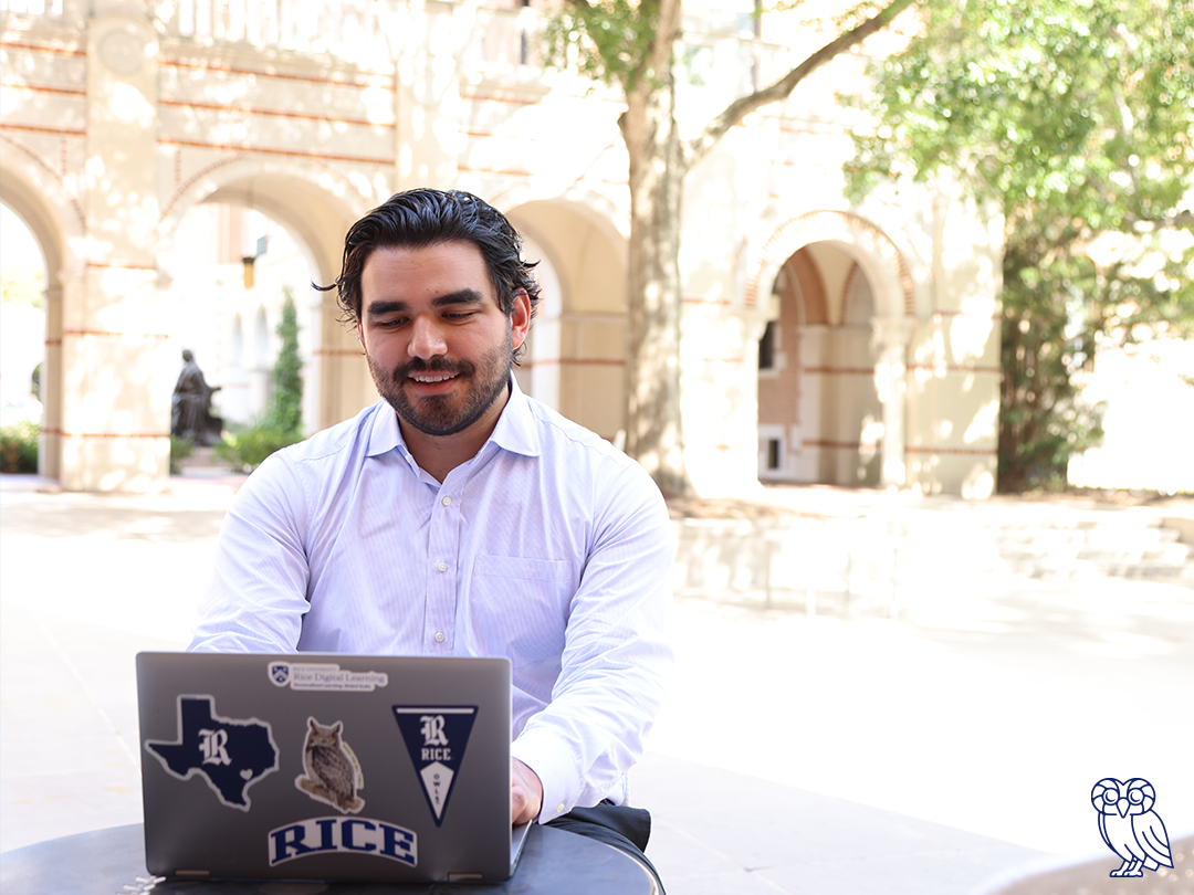 A man sitting at a desk, focused on his laptop computer, with a neutral background.